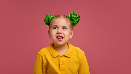 Portrait of adorable child, preschool girl wearing yellow polo shirt, with braided buns hairstyle emotionally grimacing against pink studio background. Concept of childhood, emotions, lifestyle