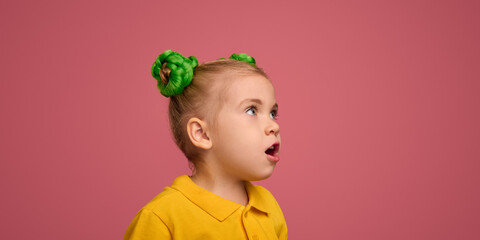 Portrait of adorable child, preschool girl wearing yellow polo shirt, with braided buns hairstyle making shocked face against pink studio background. Concept of childhood, emotions, lifestyle