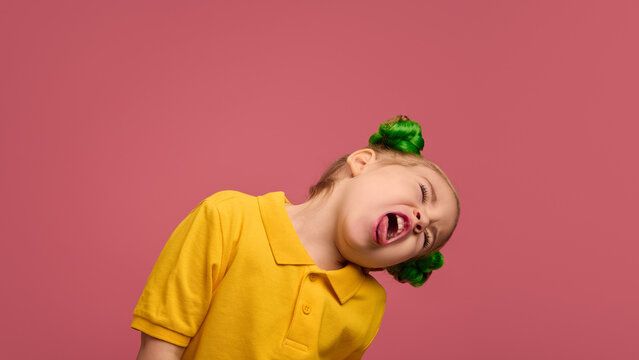 Portrait of playful child, preschool girl in yellow shirt and braided buns hairstyle emotionally making funny faces against pink studio background. Concept of childhood, emotions, lifestyle