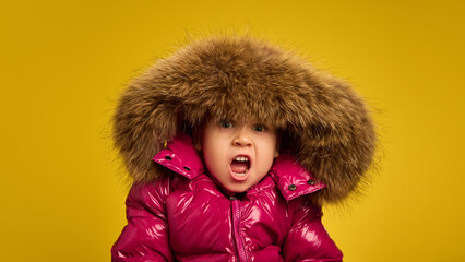 Portrait of five years old girl, emotional child in pink warm coat with large fur hood, emotionally making funny faces against yellow background. Concept of childhood, emotions, lifestyle