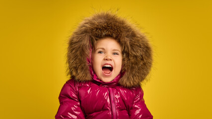 Portrait of five years old girl, emotional child in pink warm coat with large fur hood, emotionally shouting against yellow background. Concept of childhood, emotions, lifestyle