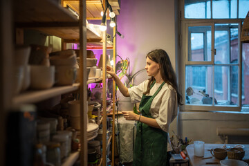 Female ceramist seller woman places completed clay items on shelves in workshop, setting up for...