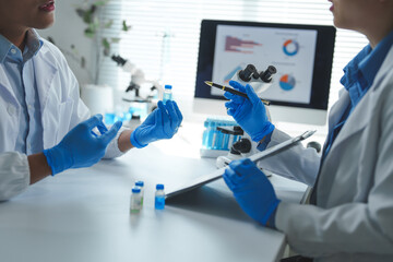 Two scientists in a laboratory discuss research findings, examining test tubes and analyzing data on a computer screen. The workspace is equipped with microscopes and lab equipment