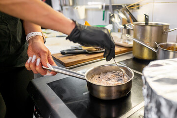 A close-up of a chef's hand stirring a sauce in a stainless steel pot on a stovetop. The scene captures a busy kitchen atmosphere, with utensils and pans in the background.