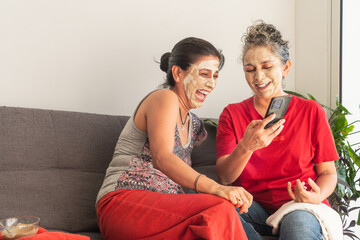 Two happy women are laughing while using smartphone and wearing face masks at home