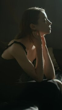 Vertical view of paranoid young woman wearing black nightdress feeling tense or nervous while sitting on couch early in morning
