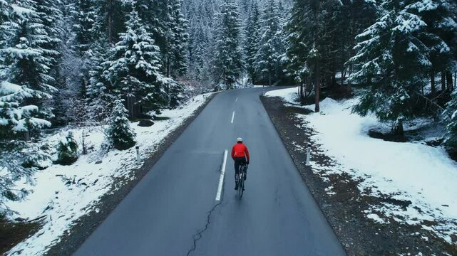 Cinematic and epic footage of cyclists on tarmac road winding between snowy winter forest, camera follow professional road cyclist on training ride in off season. Winter cycling in harsh conditions