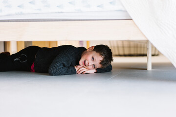 Obraz premium Boy playing under bed with a joyful expression and cozy sweater in a bright, modern bedroom