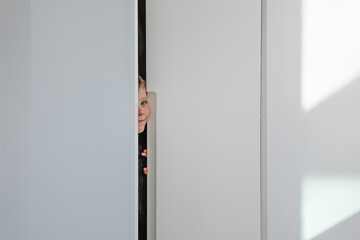Curious child peeking through a narrow gap in a white door with bright lighting illuminating the room