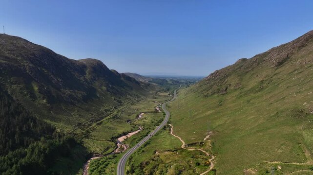 Barnesmore Gap, County Donegal, Ireland, June 2023. Drone pushes fowards in a panoramic high-angle vista through mountainous pass with a sandy river meandering next to a winding road on a sunny day.