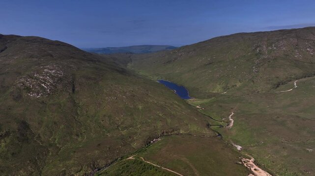 Barnesmore Gap, County Donegal, June 2023. Drone tracking right ascends behind hillslope to reveal alpine lake with winding stream system connecting down through mountainous terrain on blue sky day