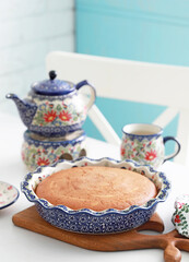 Homemade cake in ceramic form on a wooden kitchen board on a white table. A teapot on a heater and a mug in the background. Rustic still life. Selective focus.