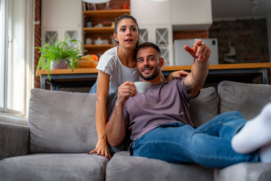 Young couple shares animated conversation about TV show as man gestures with coffee cup in hand in living room.