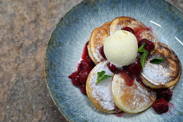 A beautifully plated dessert featuring fluffy golden pancakes dusted with powdered sugar, served with juicy red cherries in syrup.