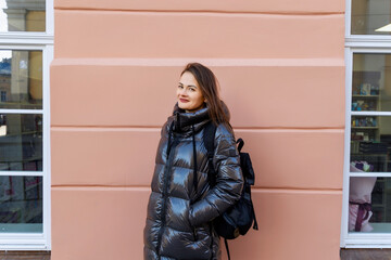 Woman in shiny jacket standing near peach wall, smiling woman in shiny jacket with backpack near peach wall. © Iryna