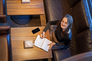 Woman writing in journal at cafe table. A woman in glasses looks up while writing in a journal at a wooden cafe table, with a smartphone and tissue box nearby.