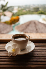 Morning hot Americano in a white mug with frangipani flower. Black coffee on a bamboo table. A cup of strong coffee in the morning on vacation