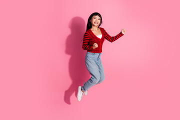 Cheerful young woman in a red striped cardigan enjoying joyful jump in front of a pink background