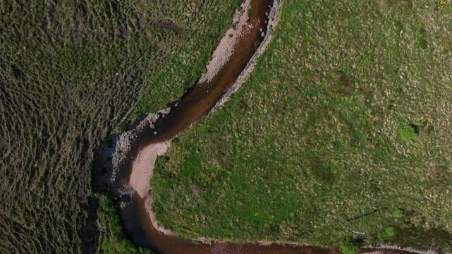 Barnesmore Gap, County Donegal, June 2023. Drone top down pan follows slow moving river with built up sandy riverbanks moving along sloped grassy bogland hills
