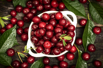 A bowl brimming with glistening, ripe red cherries sits amidst lush green leaves on a dark rustic surface. The vibrant colors and natural setting create a tempting and visually appealing image