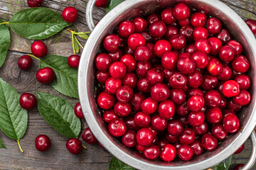 A bowl brimming with glistening, ripe red cherries sits amidst lush green leaves on a dark rustic surface. The vibrant colors and natural setting create a tempting and visually appealing image