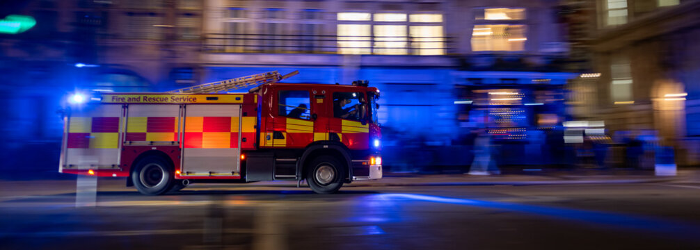 A fire truck speeds through the city at night with emergency lights flashing, responding to an urgent call. The motion blur effect emphasizes speed and urgency. Firefighters are visible inside.