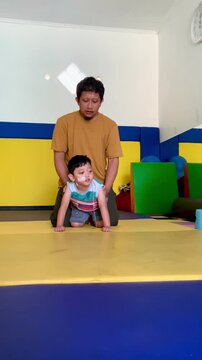 A father trains a toddler who lives with cerebral palsy and installs an NGT nasogastric tube in his nose to learn to crawl in the physiotherapy room.