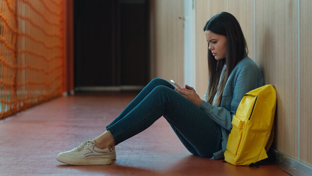 Sad upset failure stress pensive thinking girl female woman student pupil education studying sitting floor in school university college corridor using mobile phone smartphone online app social media