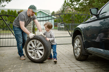 Cute little boy helping his father to change car wheels at their backyard. Father teaching his little son to use tools.