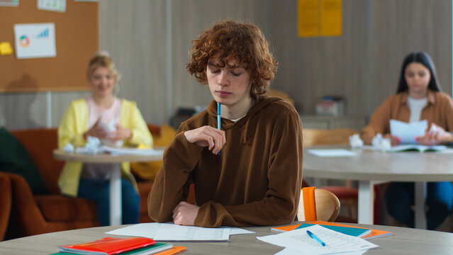 High school bullying upset stressed lonely Caucasian guy teen male boy schoolboy nerd student teenager writing task class desk university college children students mock bully throwing papers balls