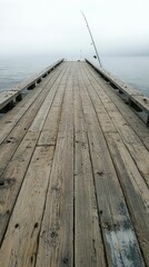 Fototapeta premium Wooden pier extends to foggy water; fishing rod rests.