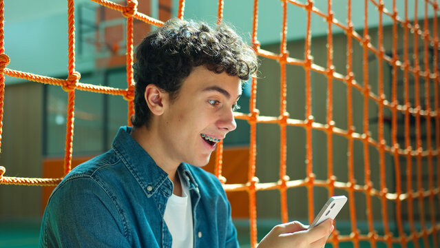 Shocked amazed wonder Caucasian teen boy student guy teenager man male son pupil in university school corridor sport hall sitting on floor shock mobile phone stunned smartphone social media addict