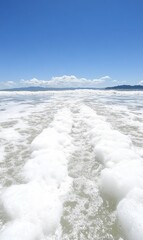 Obraz premium Sea foam trail on sandy beach with distant islands under clear blue sky, ideal for travel ads