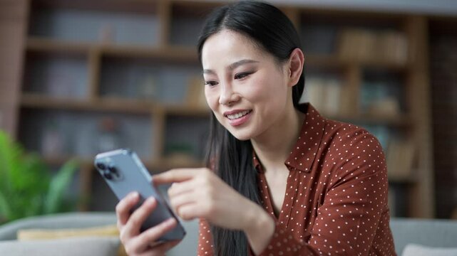Happy young asian woman using mobile phone while sitting on sofa in living room at home. Smiling female browses social media, makes purchases, chats with friend, writes or reads messages. Close up