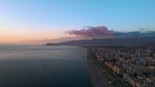 Cinematic drone view of numerous high rise buildings on the shore of the sea with a beach and a road along the coastline of Mahmutlar, Alanya, Turkey, with epic pink clouds and mountains in the backgr