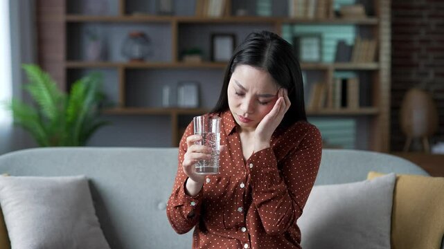 Young asian woman suffering from headache while sitting on sofa in living room at home. Sad female massages her temples with her hand while holding a glass of water. She feels pain. Migraine. Close up