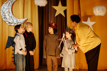 Group of children dressed in costumes, preparing for a school play with teacher supervising their performance. Stars and moon decorations hang in the background