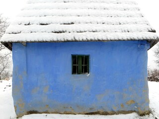 Old snow-covered blue mud house with rustic wooden roof in a winter landscape, highlighting traditional architecture in a peaceful snowy countryside setting in historic Maramures, Romania