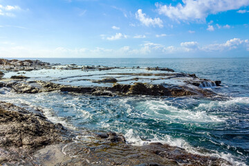 A serene coastal scene featuring rocky formations, gentle ocean waves, and a bright blue sky with scattered clouds.