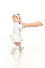 Adorable baby, girl in white outfit and cute hair accessory, holding hands with her mother and walking against white studio background. Concept of childhood, motherhood, care. Copy space for ad