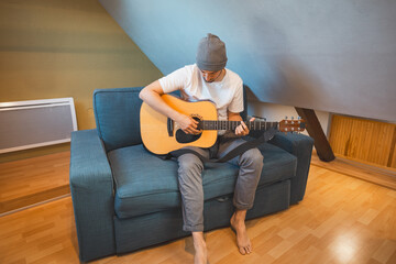 Musician 26 years old sits barefoot on a cozy blue couch in an attic room, strumming an acoustic...