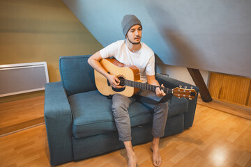Musician 26 years old sits barefoot on a cozy blue couch in an attic room, strumming an acoustic...