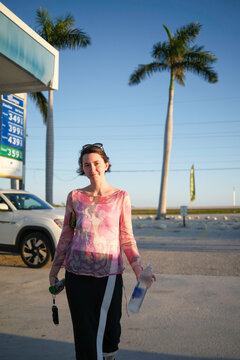 Woman stands outside a gas station holding water bottle and keys, palm trees in the background, Florida Keys, USA