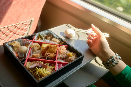 Bento box with assorted dishes on a tray in a sunlit train, hand holding chopsticks, near Kanazawa, Japan