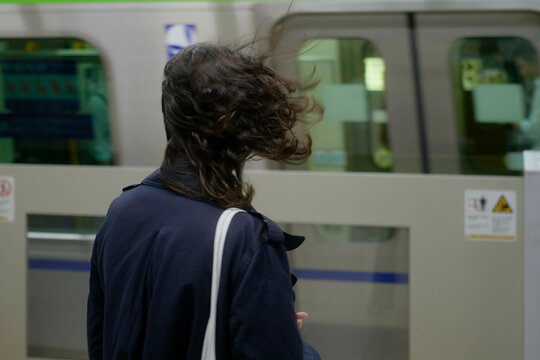 Woman with windblown hair stands at a subway platform as a train passes by. Tokyo, Japan