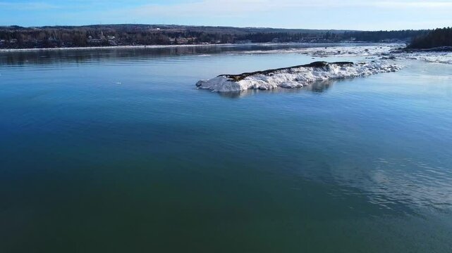 Winter drone approach toward an ice‑covered tidal rock in the calm Baie de Mitis along the St. Lawrence River near M&eacute;tis‑sur‑Mer, Quebec, Canada