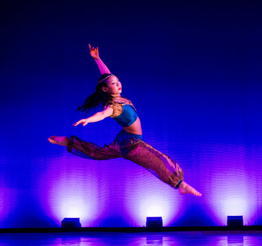 Dancer in mid-air leap wearing colorful costume against vibrant blue-lit background. The Nutcracker, Bainbridge Island, USA