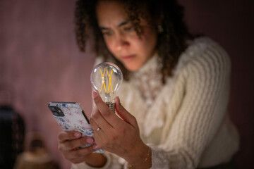 A woman holds a clear light bulb and looks at her smartphone with focused attention while researching. Netherlands