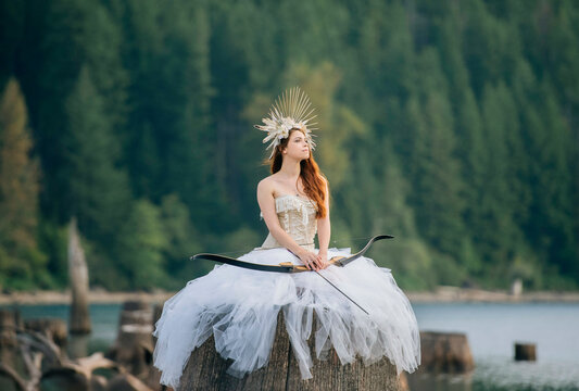 Woman in a white dress and headpiece sits on rock with bow near a lake in a forest. Rattlesnake Lake, Puget Sound, WA, USA