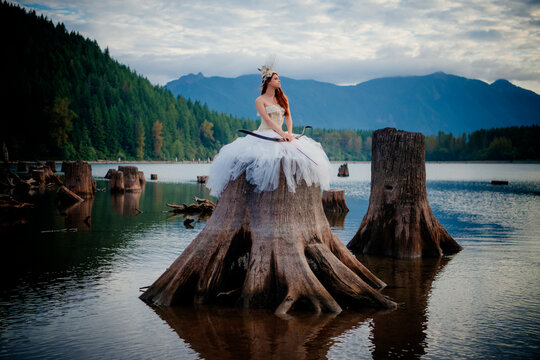 Woman in white gown with bow and arrow sits on massive tree stump by tranquil lake with forest and mountains. Rattlesnake Lake, Puget Sound, WA, USA